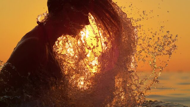 SLOW MOTION, SILHOUETTE, LENS FLARE, COPY SPACE, DOF: Cinematic shot of young woman pulling her head out of the ocean and flipping her hair back, spraying glassy droplets into the sunny evening sky.