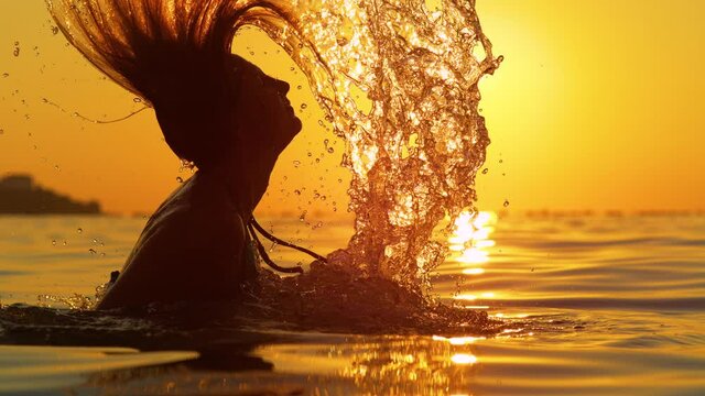 SLOW MOTION, SILHOUETTE, LENS FLARE, DOF: Golden summer sunset shines on the beautiful young woman flipping her wet hair back after pulling it out of the glassy ocean. Female tourist on vacation.