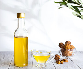 Walnut oil and a bunch of nuts on a white wooden table. Modern still life with oil bottle and glass bowl with walnuts, olive branch and copy space.