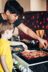 Father and son sprinkle grated cheese on pizza. Preparing for the Mothers Day holiday. Asian father and mestizo son prepare food in the kitchen