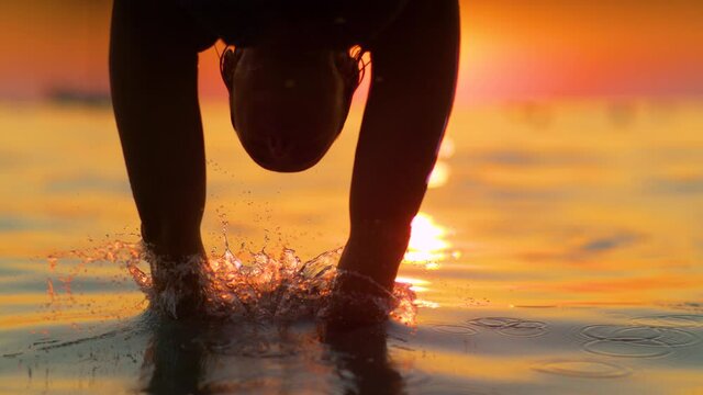 SLOW MOTION, CLOSE UP, LENS FLARE, SILHOUETTE, DOF: Female traveler jumps into the refreshing sea water on a sunny summer morning. Golden sunrise shines on young woman diving into the Adriatic sea.