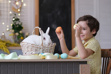 Happy easter. Child boy in bunny ears plays with decorated eggs and hare sits in a wicker basket on the kitchen table. soft focus