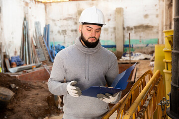 Naklejka na ściany i meble Young bearded foreman making task list during works in building under construction