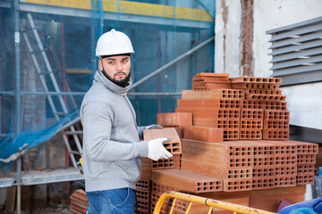 Young bearded bricklayer taking red bricks from stack in building under construction
