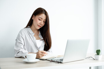 Asian female college students study online at home, enjoy extra lessons, watch videos, and take notes from a digital laptop, do homework by looking at the screen while sitting at a desk at home