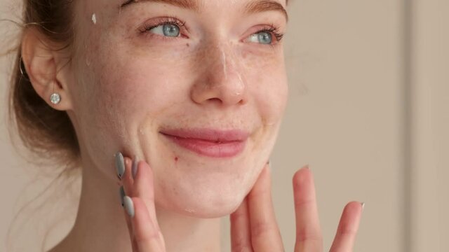 Close up photo of a red haired woman applying cream on her freckled face and smile