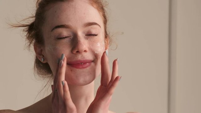 Close up photo of a ginger woman with freckles massaging her face with cream and smile at camera with bare shoulders