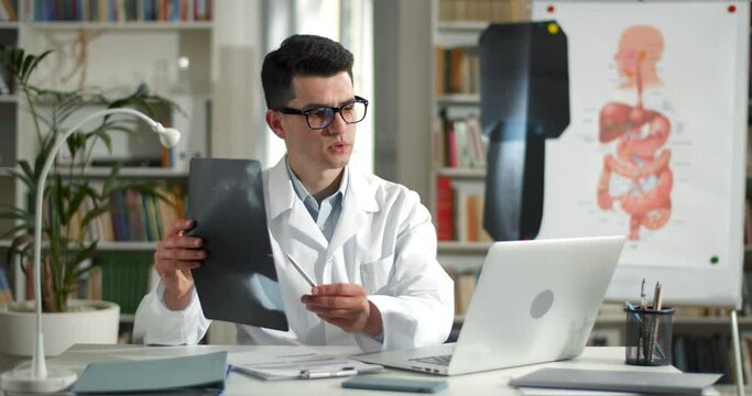 Male doctor holding x ray photo while having online medical consultation in office. Man in glasses and white rob using laptop while communicating with patient and sitting at table.