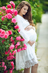 Smiling pregnant woman resting in near roses. Posing outdoors. Motherhood. High quality photo.