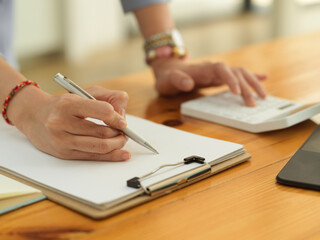 Female hand with pen writing on blank paper on clipboard and using calculator on wooden office desk