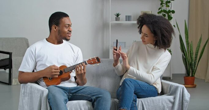 Afro american man musician sitting on couch singing playing small hawaiian guitar musical instrument girl curly woman using mobile phone helping boyfriend record video music lesson taking photos