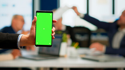 Businesswoman holding smartphone with green screen while diverse team working on background, businesspeople analysing financial statistics, Multiethnic coworkers planning project on chroma key display