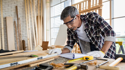 Aged Asian man reading manual while making timber furniture in joinery. Older retired people hobby and  DIY in free time  concept - obrazy, fototapety, plakaty