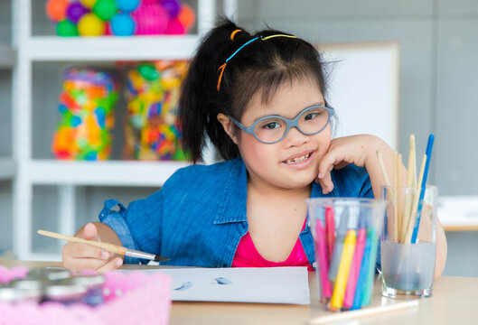 Young Asian down syndrome little girl sitting at desk with colors prepare for painting. She widely smiles with bright eyes and happiness and looking to camera. Concept for education for disabled kid