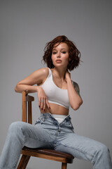 A young beautiful woman with plump lips and short wavy red hair poses in a white T-shirt on a gray...