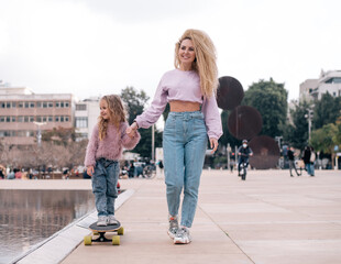 Mother and daughter playing on skateboard. Woman with unusual, lush and long hair. Small girl and woman smiles and happy. Same colour  of clothes, get fun outdoor in weekend. Small kid study skating. 