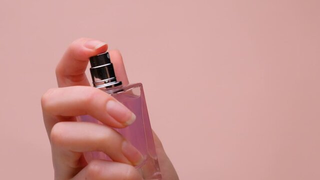 A woman's hand holds a bottle of pink perfume and presses a finger on the cap. Splashes fly out. Beige background