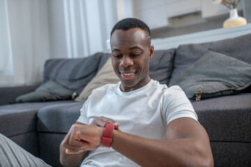 Dark-skinned young guy sitting on the floor and looking at his smartwatch