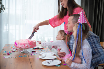 cheerful Caucasian family sitting at the table at home and celebrating a birthday. mom, dad and two daughters. A woman is cutting a large cake into pieces.