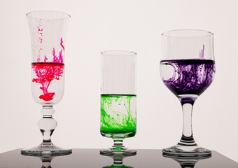 Backlit and reflecting surface scene of different food colouring dye droplets falling down into water inside of glasses.
