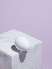 Geometric composition with marble cube and white egg on the edge against pastel violet background. Creative Easter holiday concept. Abstract isometric food layout with trend summer shadows.