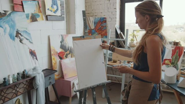 Young female artist holding palette and putting blank canvas on easel while working in studio