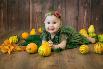 Fototapeta premium Cute little girl up to 1 year old in a green cute dress and a floral wreath with small yellow and orange pumpkins on a brown wooden background 
