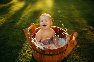 baby splashes in a basin in nature on a green tvava, at sunset. Good mood, child's smile, happiness from swimming in water, water splashes fly, good summer day