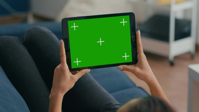 Close up of woman holding in horizontal mode tablet computer with mock up green screen chroma key display lying on sofa. Freelancer using isolated touchscreen device for social networks browsing