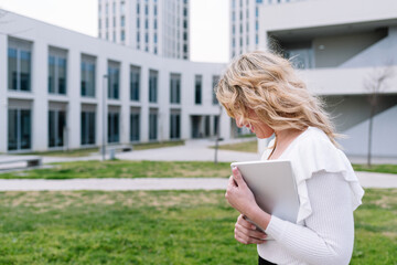 Elegant woman walking with laptop in hand with office buildings in background. The blonde-haired executive moves from place to place down the street.