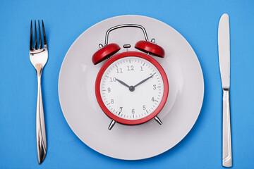 Red alarm clock lies on plate next to fork and knife
