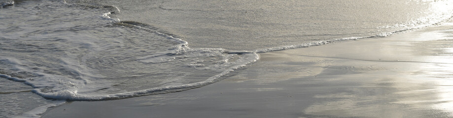 Vagues s'étalant sur une plage mouillée, en contre-jour un jour ensoleillé, format panoramique