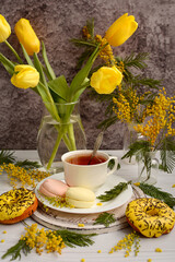 Vertical composition of yellow flowers and donuts and a white cup with tea on a light wooden background