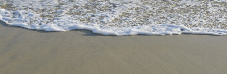 Écume blanche d'une vague sur la moitié du sable fin d'une plage ensoleillée, vue de face, formant une ligne horizontale, format bannière