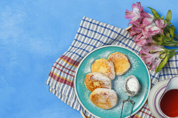 A plate of cheesecakes with powdered sugar, a jug and flowers on a blue surface, horizontally with space