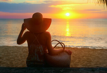 Vacation Summer Holiday Concept. Woman in dress with summer bag watching sunset on beach.