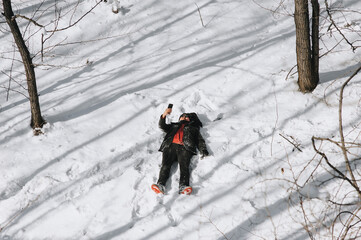 A wanderer, a tourist with a backpack lies on the snow in the forest and photographs the beautiful nature and himself on a mobile phone.