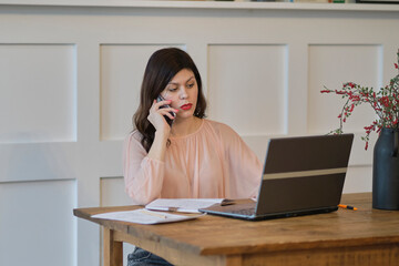 Businesswoman talking on mobile phone working on laptop in cozy home office. free space startup...