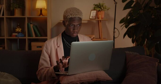 Crop view of young stylish man typing and using laptop while sitting on couch. Serious afro american guy with dyed hair browsing internet at home. Concept of leisure.