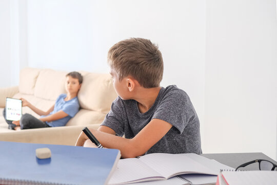 Boy doing homework with books notebooks and other boy using digital tablet for online lessons. Brothers at home talking about homework tasks