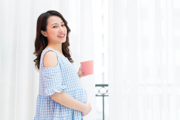 Beautiful woman holding her stomach and holding a cup of milk stands by the window