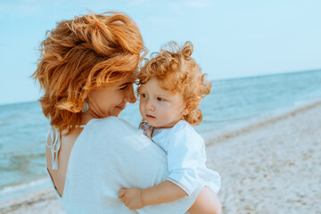Mom is holding and smiling a little daughter 1 year old with red curly hair in light clothes in the...