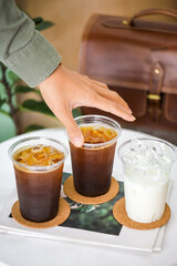 A hand is going to pick up a glass of amicano coffee drink form the table which have 2 glasses of coffee drink and a glass of iced fresh milk.