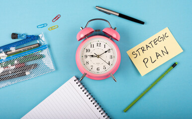 Fototapeta na wymiar A table clock with words Strategic plan Written on a yellow sticky note and Pen with other elements on a blue background