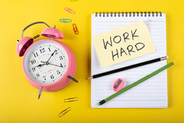 A table clock with Work hard words Written on a sticky note with notepad pencil and other elements on a Yellow background