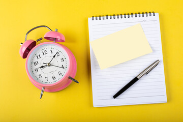Table clock with notepad sticky note and a pen on a yellow background flat lay shot.