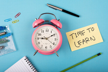 Fototapeta na wymiar A pink color table clock with Time to learn words Written on a yellow sticky note and Pen with other elements on a blue background