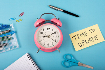 A pink color table clock with Time to update words Written on a yellow sticky note and Scissor with other elements on a blue background flat lay shot