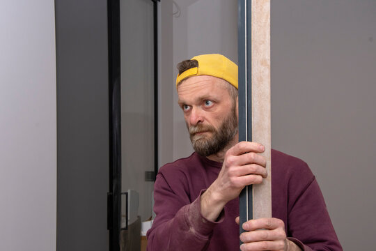 A 45-50-year-old Male Construction Worker In A Yellow Cap Holds A Door Trim Or Skirting Board In His Hands. Concept: Illegal Work Abroad, Interior Decoration, Ad Work.