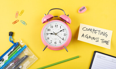 Fototapeta na wymiar A table clock with Competing against time words Written on a sticky note with other elements on a Yellow background flatlay shot